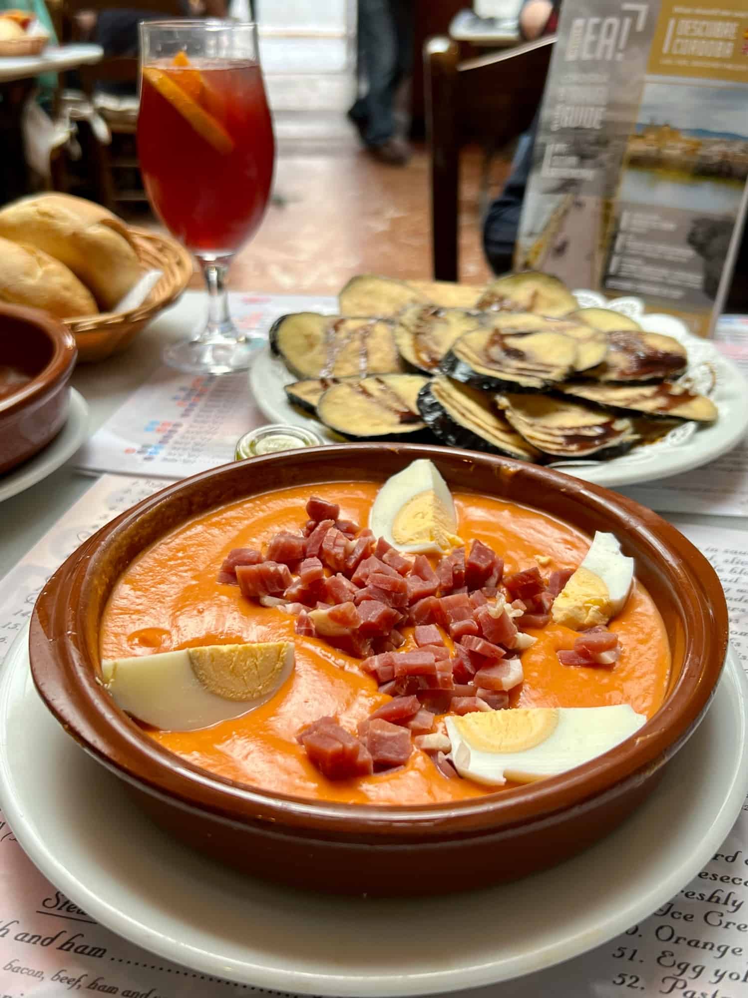 A bowl of chilled salmorejo soup with hard boiled eggs and ham with a plate of fried aubergines in honey in the background.