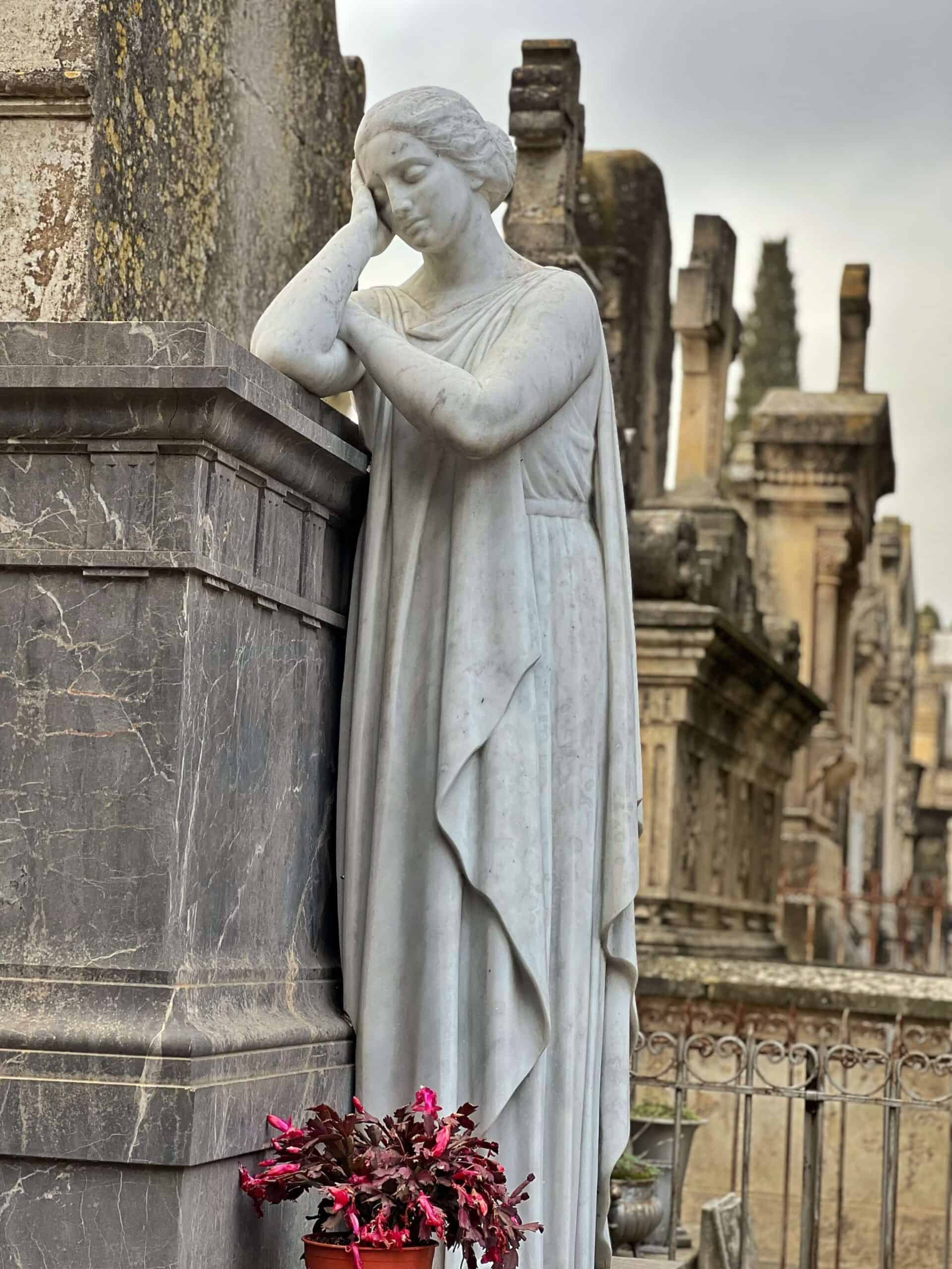 A white statue of a woman grieving beside a mausoleum in a cemetery