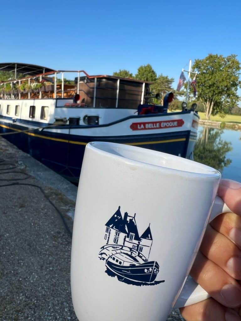 holding a white coffee mug with a drawing of the barge in front of the barge