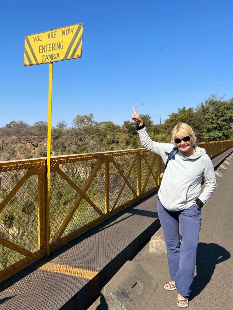 Lady wearing grey fleece, navy trousers and sunglasses standing on the old bridge at Victoria Falls pointing to a yellow sign 