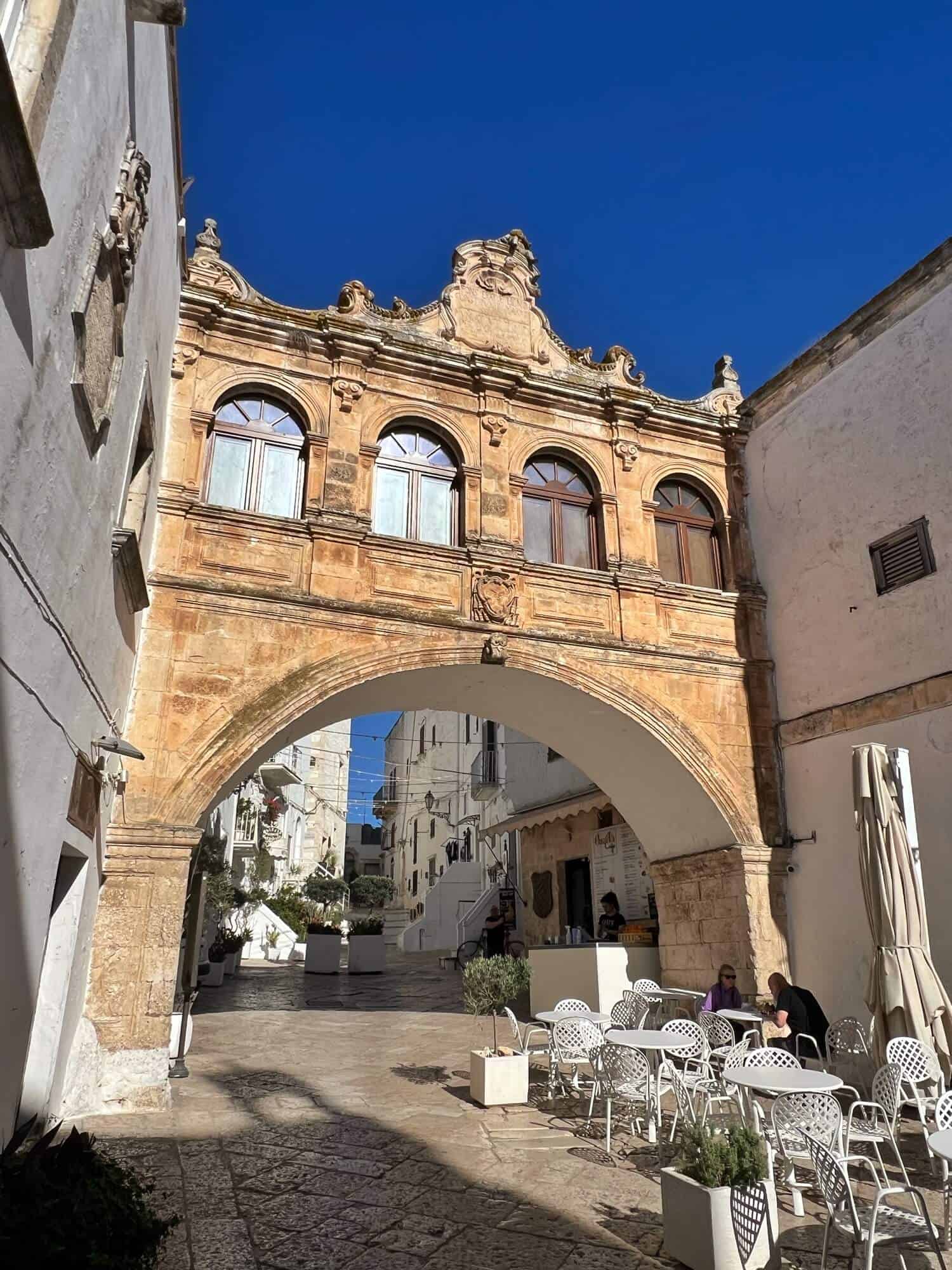White chairs and tables beneath a blue sky and a historic archway in Ostuni 
